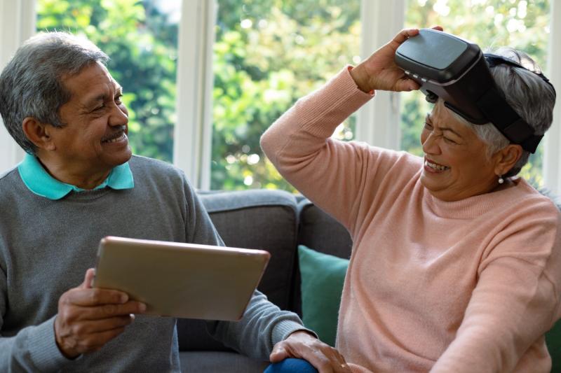 An old man holding an ipad smiling to an old lady holding an VR device